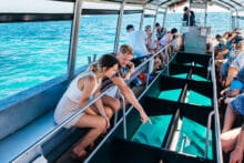 Passengers looking through the glass panels of a glass-bottom boat over clear coral reef water.