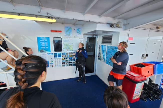 Dive instructor leading a morning reef-site briefing with guests on OceanQuest.
