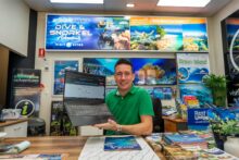 Clint Carroll of Cairns Dive Adventures displaying new AI-powered Reef Tour Matchmaker quiz on laptop in Cairns office surrounded by reef tour posters