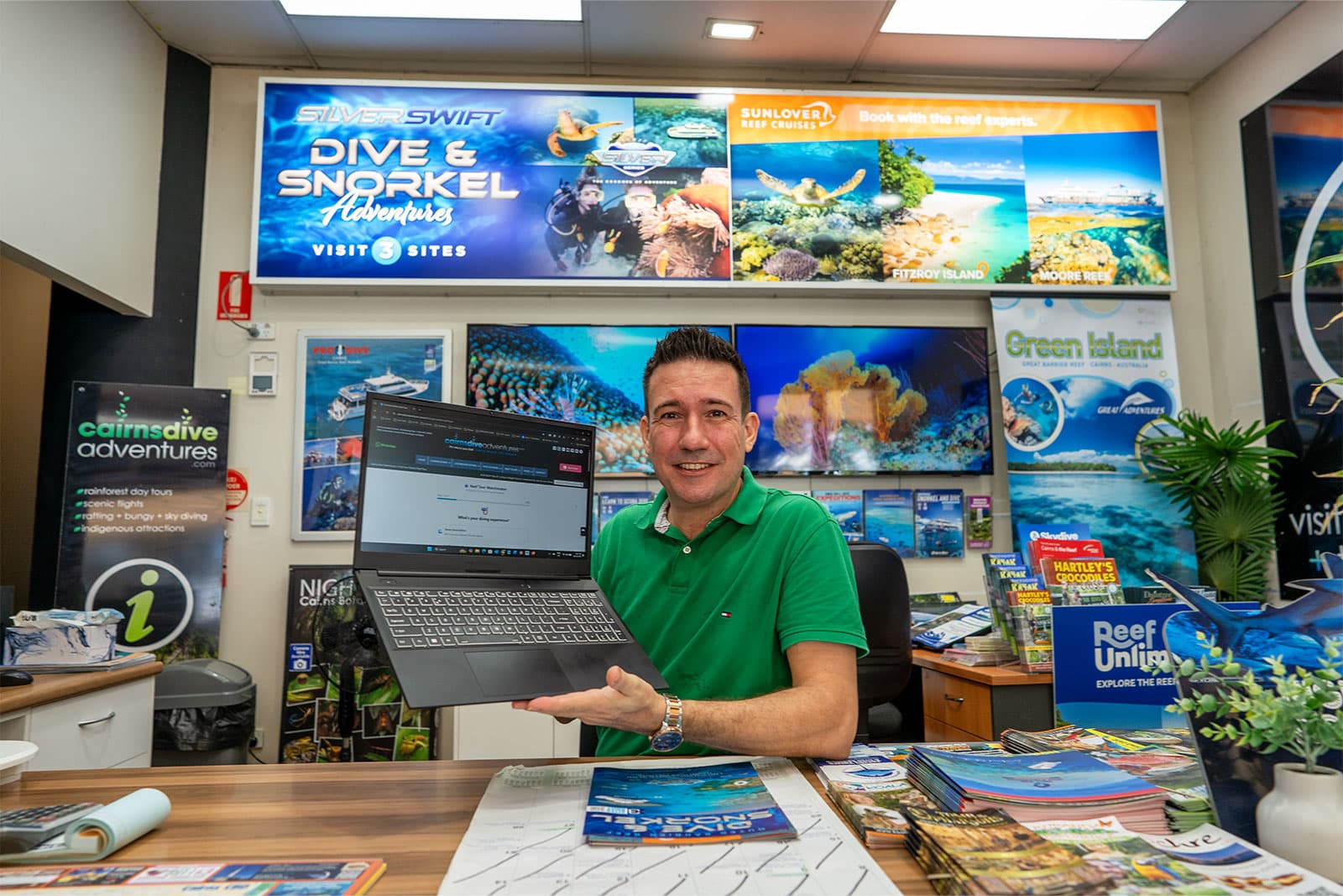 Clint Carroll of Cairns Dive Adventures displaying new AI-powered Reef Tour Matchmaker quiz on laptop in Cairns office surrounded by reef tour posters